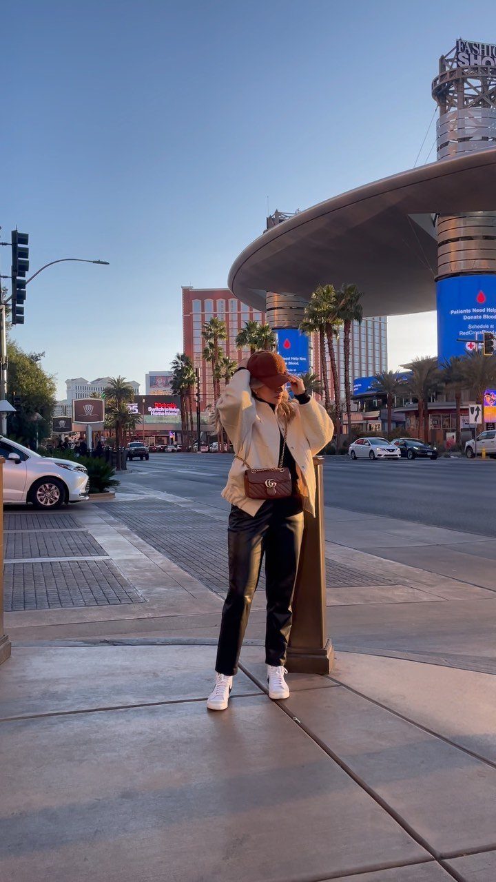 A woman in black leather pants and a cream jacket wearing white sneakers on a city street.