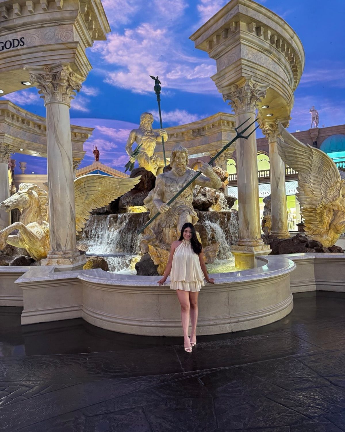 Woman in a cream mini dress and sandals sitting on an ornate fountain edge.