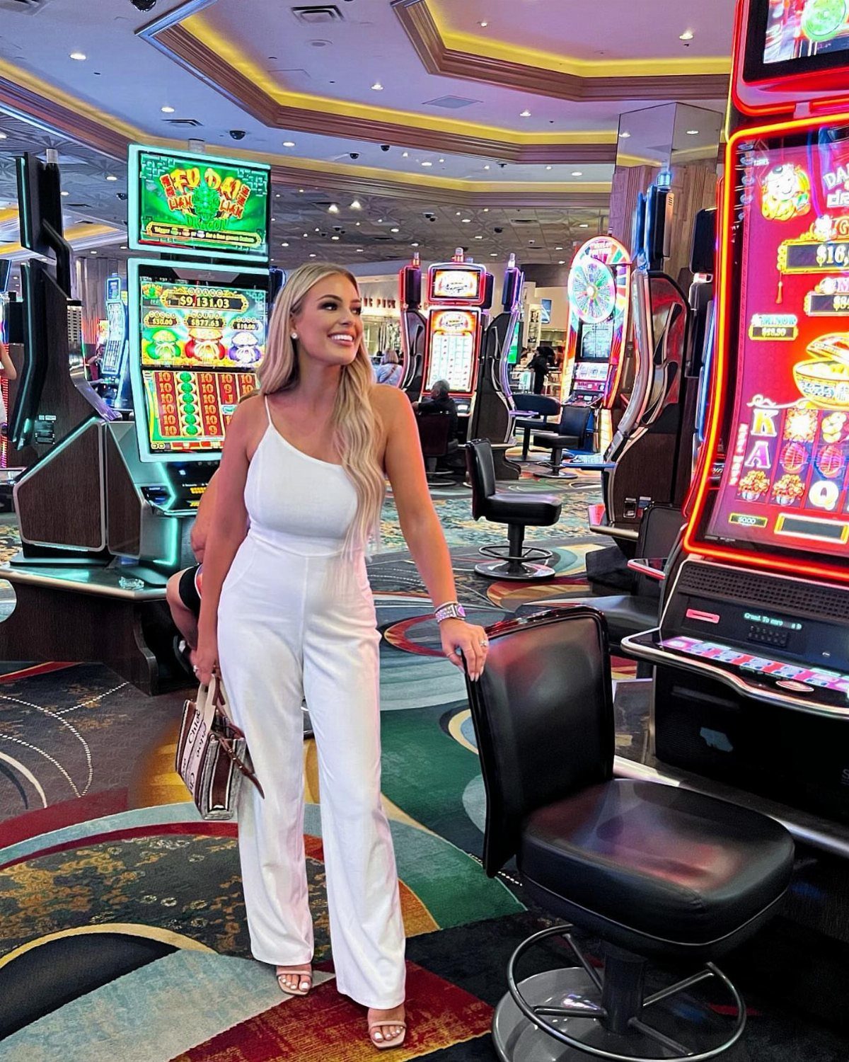 Blonde woman in a white jumpsuit smiling in a brightly lit casino among slot machines.
