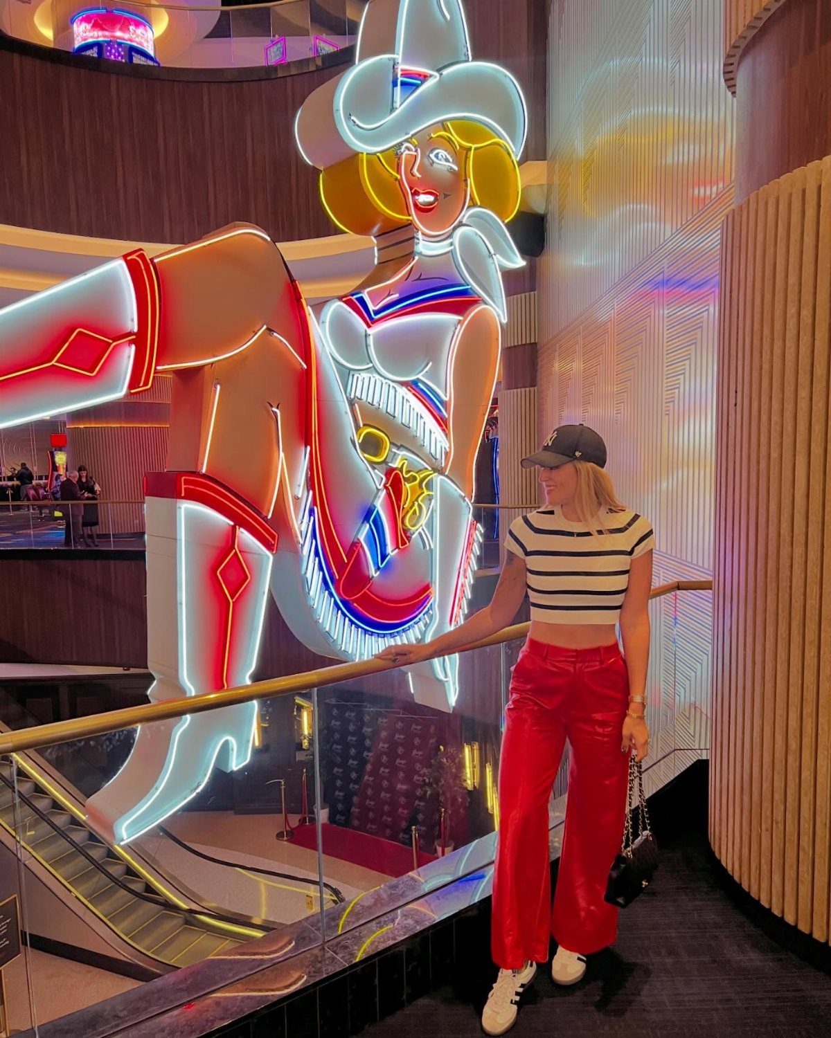 Woman in striped crop top and red wide-leg pants standing near a large neon sign.