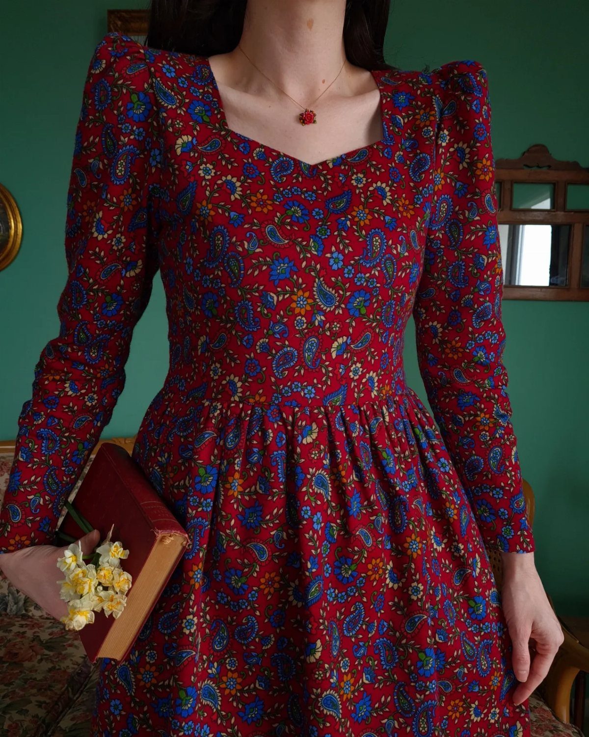 Person wearing a red floral puff-sleeve dress holding a book and yellow flowers.