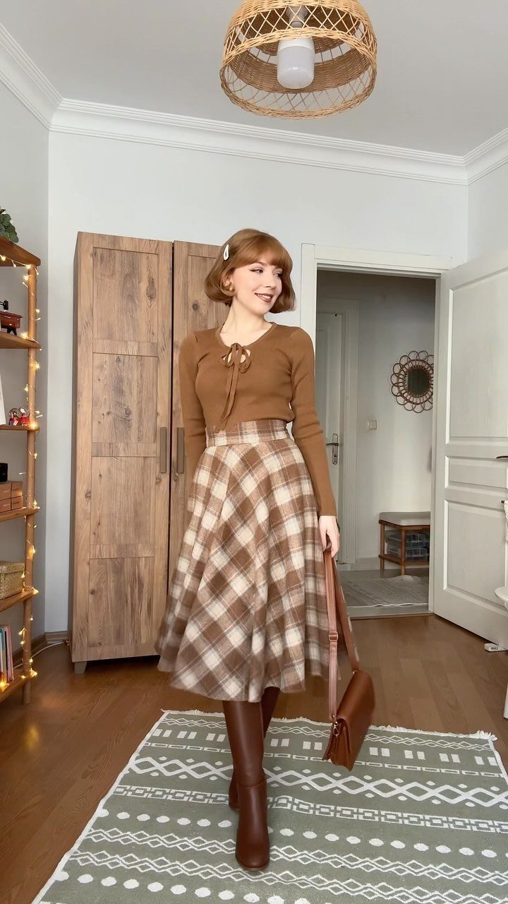 A woman smiling in a brown long-sleeved top, plaid midi skirt, and tall brown boots.