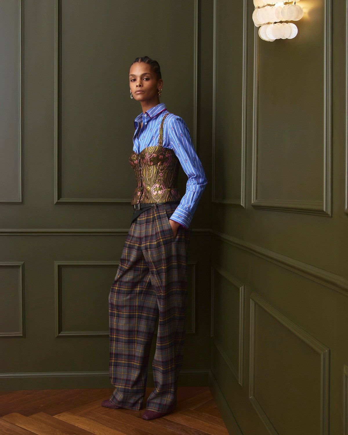 Woman wearing a blue patterned shirt, floral corset, and plaid wide-leg trousers against green walls.