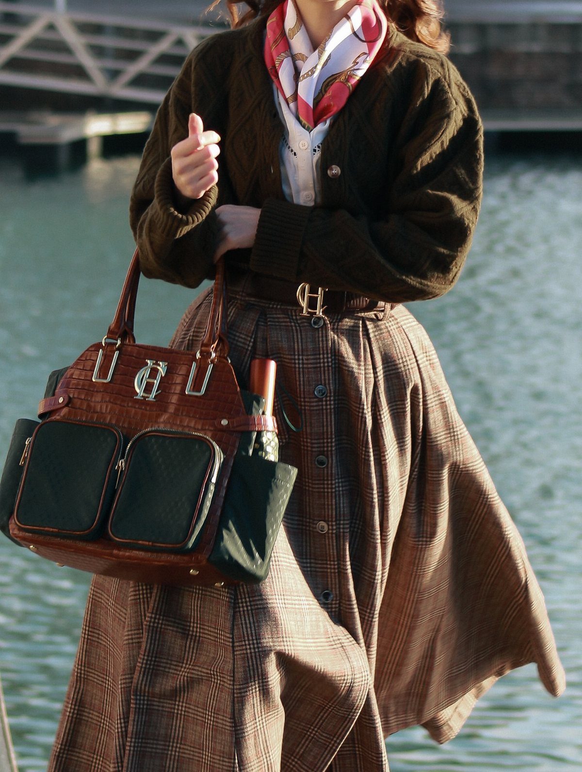 Person wearing a brown plaid swing skirt, dark cardigan, and patterned scarf by the water.