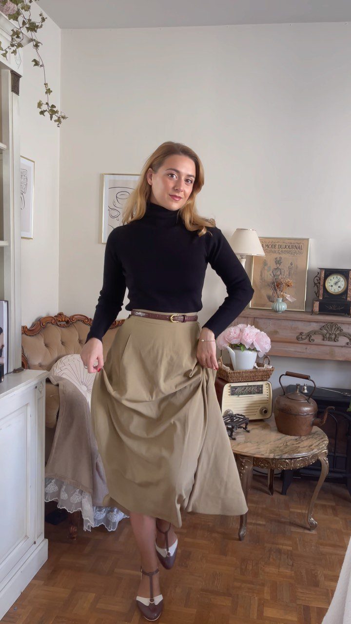 A woman wearing a black turtleneck and tan midi skirt posing in a vintage-style room.