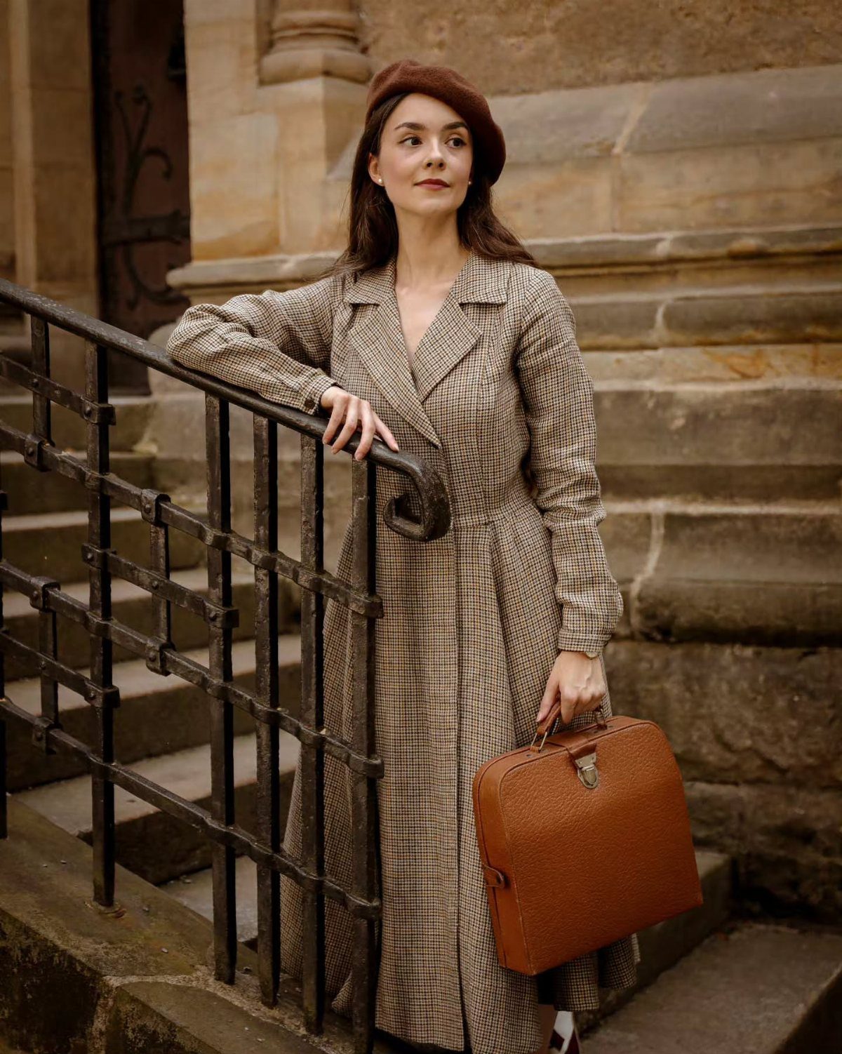 A woman in a plaid coat and beret standing on stone stairs holding a briefcase.