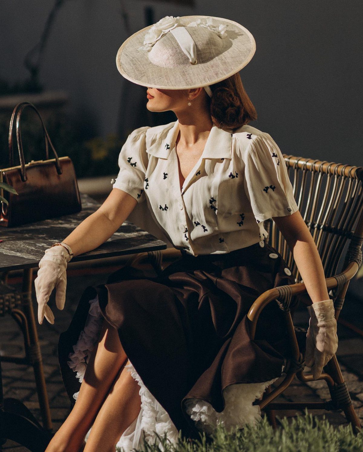 Woman in vintage cream blouse, dark skirt, lace gloves, and wide-brimmed hat sitting outdoors.