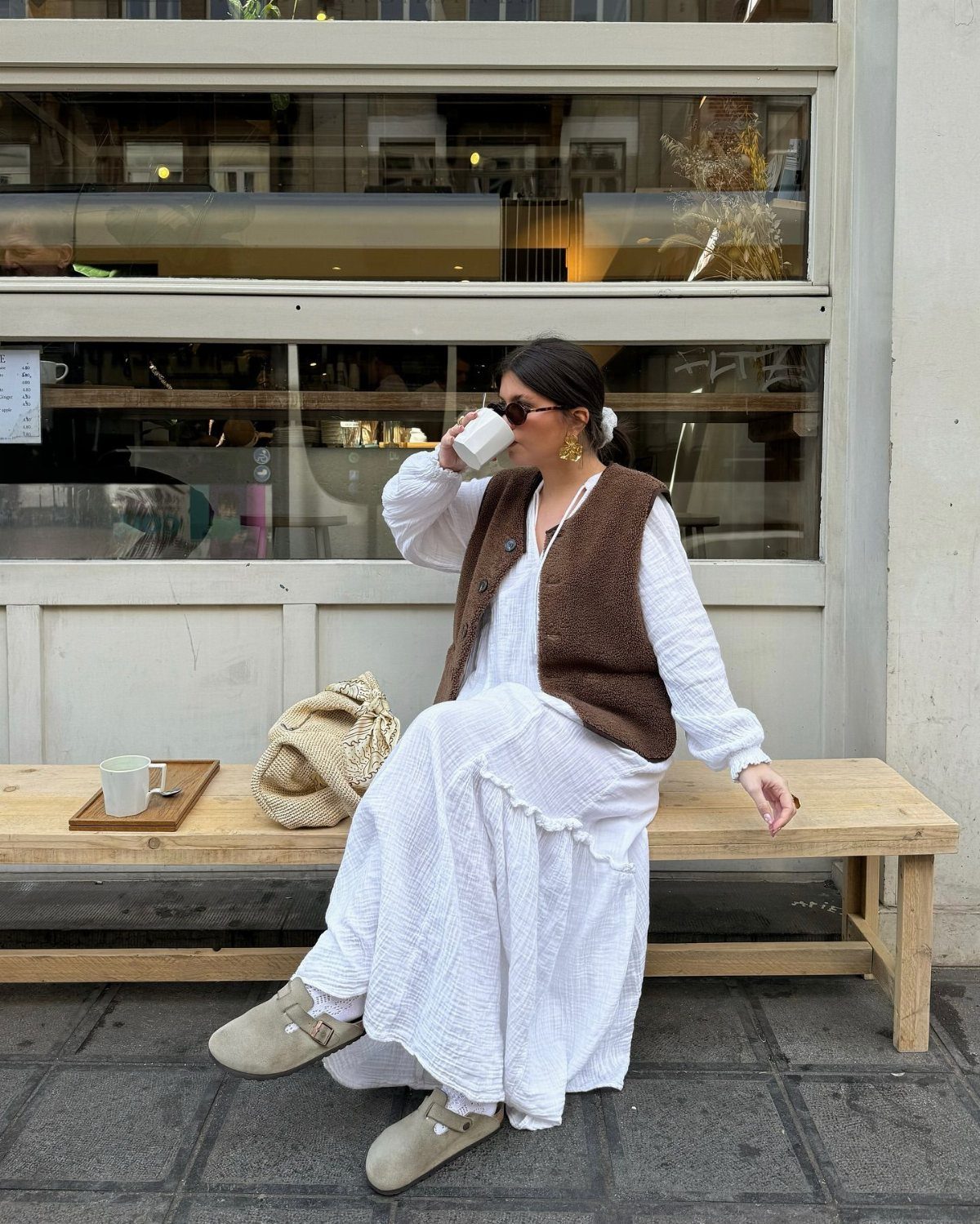 A woman in a white cotton maxi dress and brown vest sitting on a bench.