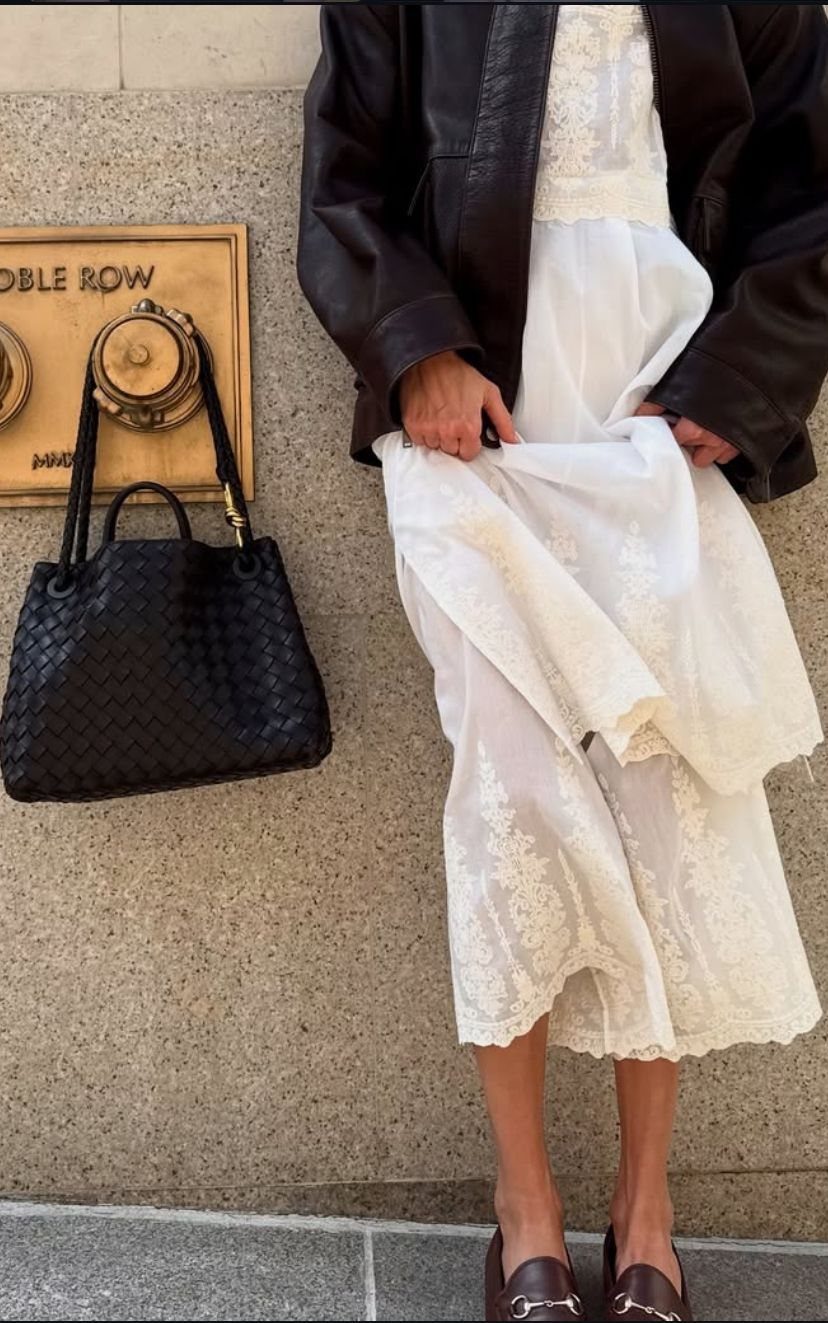 A person wearing a white lace midi dress and a dark brown leather jacket standing outdoors.