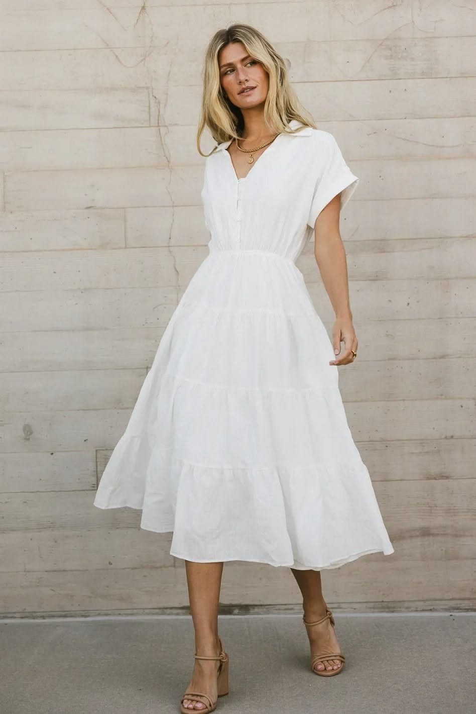 A woman posing in a white cotton midi dress with a tiered skirt against a stone wall.