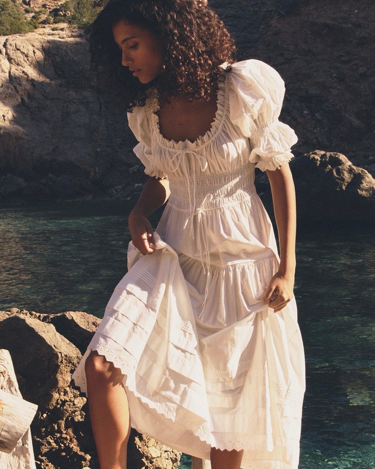 A woman with curly hair wearing a white puff-sleeve cotton sundress by the ocean.