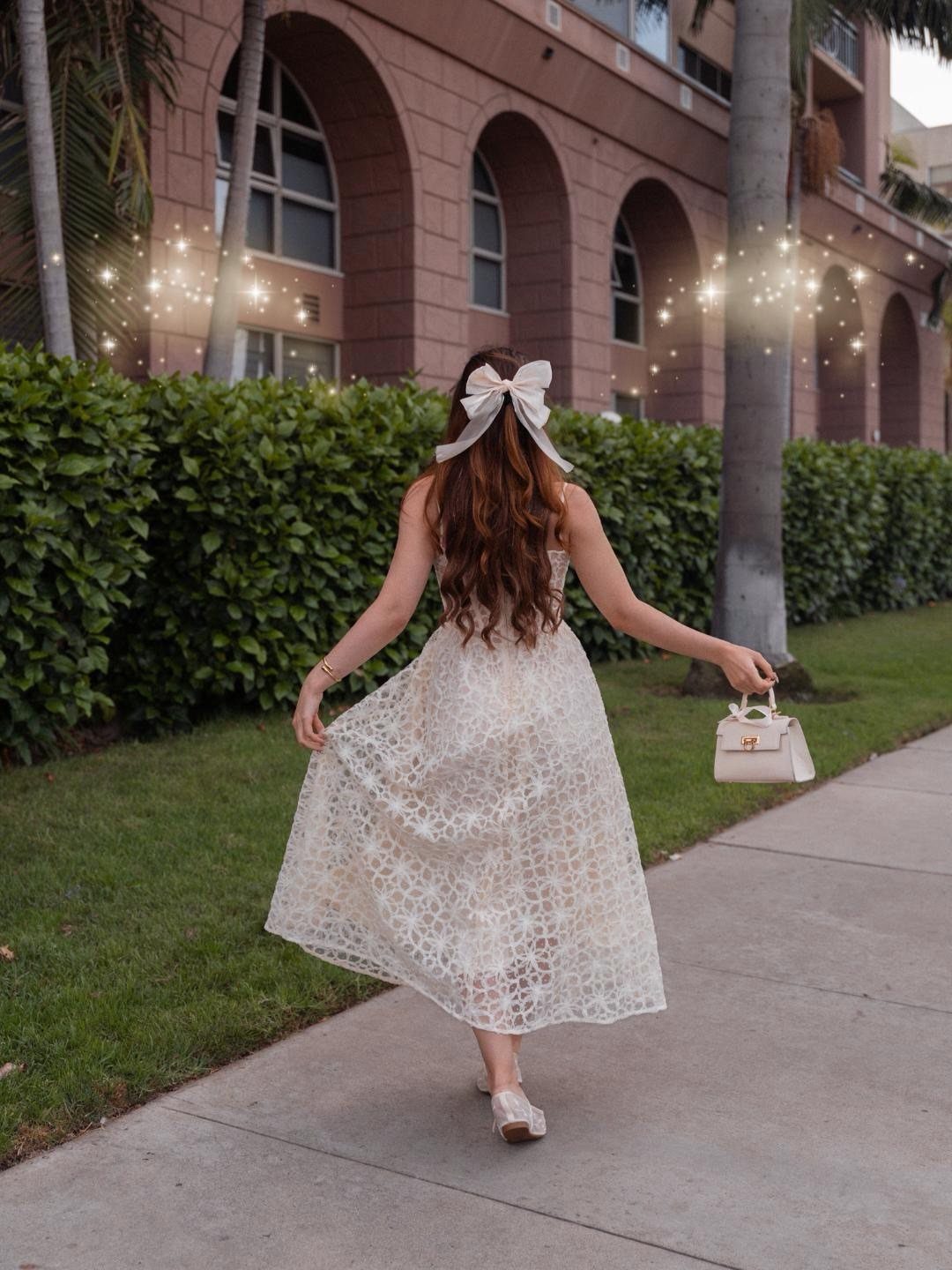 Woman in a white lace midi dress walking along a sidewalk with a white bow in her hair.