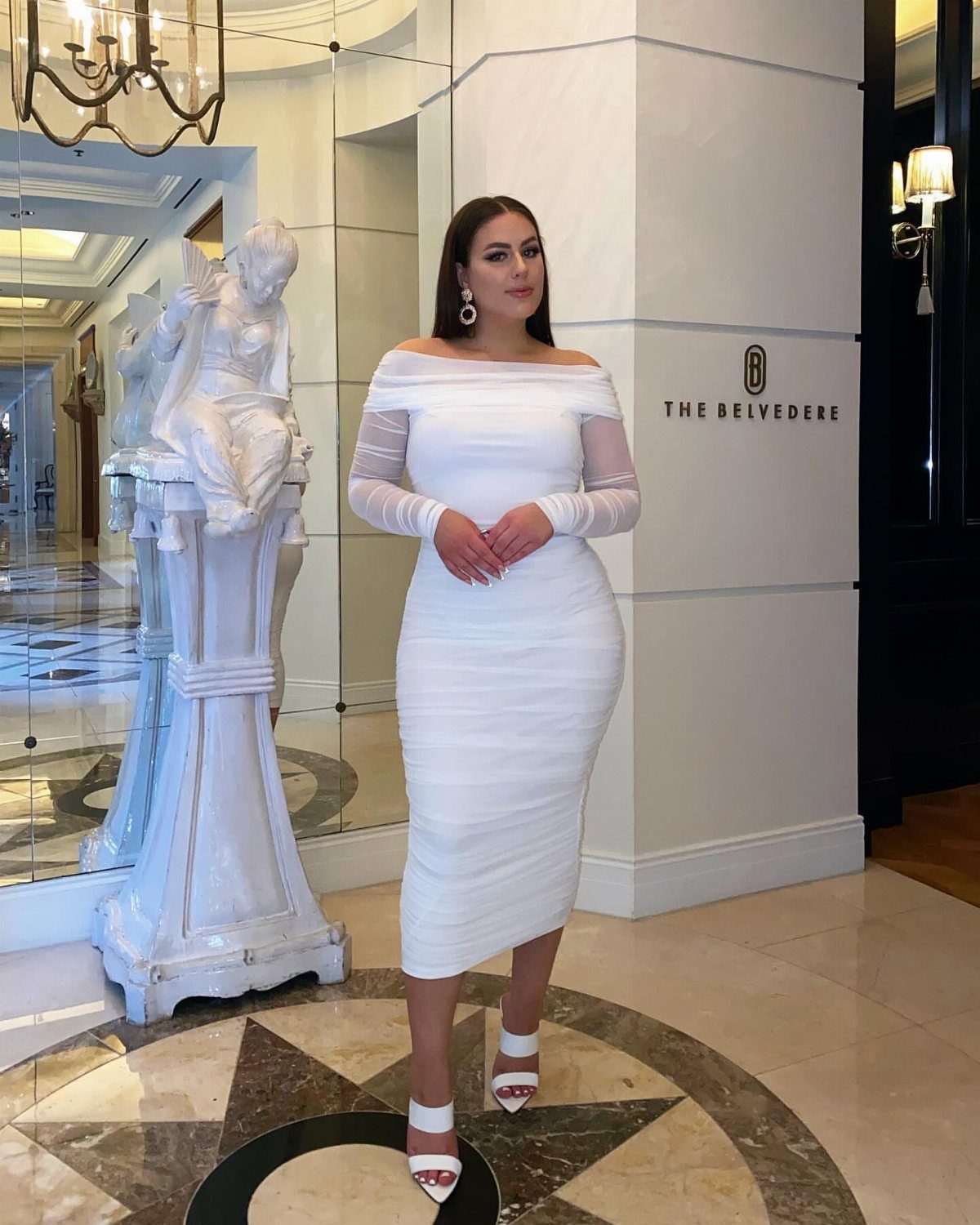A woman posing in a white off-the-shoulder midi dress inside an elegant marble lobby.