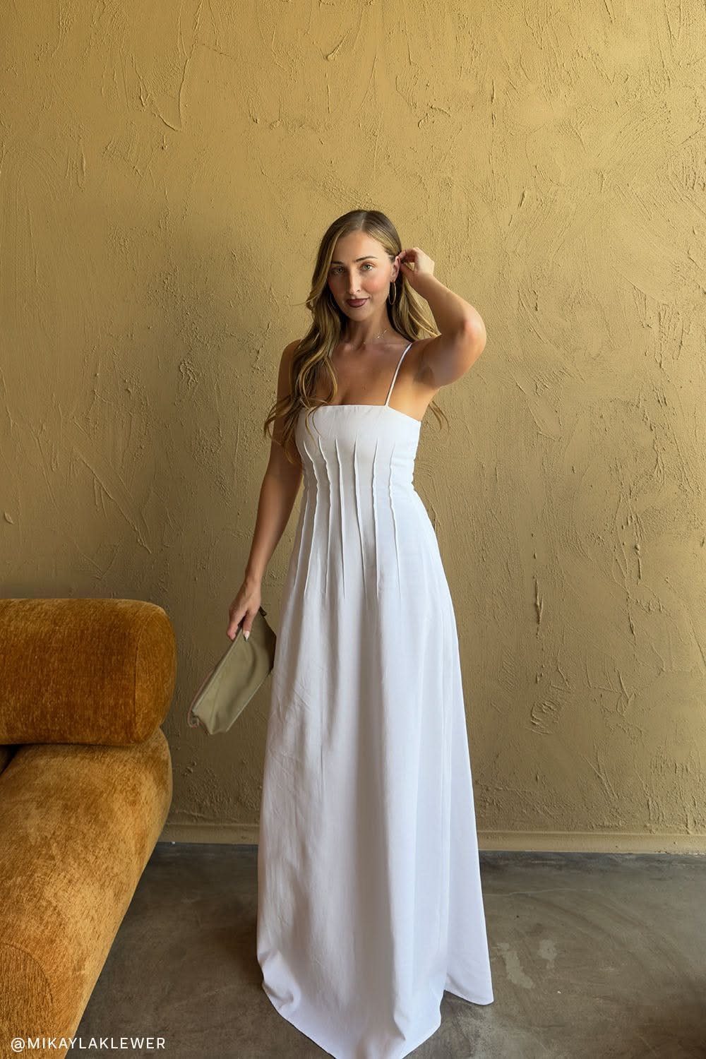 A woman in a white spaghetti strap maxi dress posing against a tan wall.