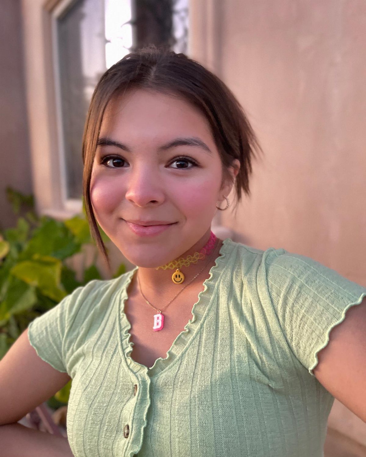A smiling young woman wearing a pink beaded choker and a light green ribbed top.