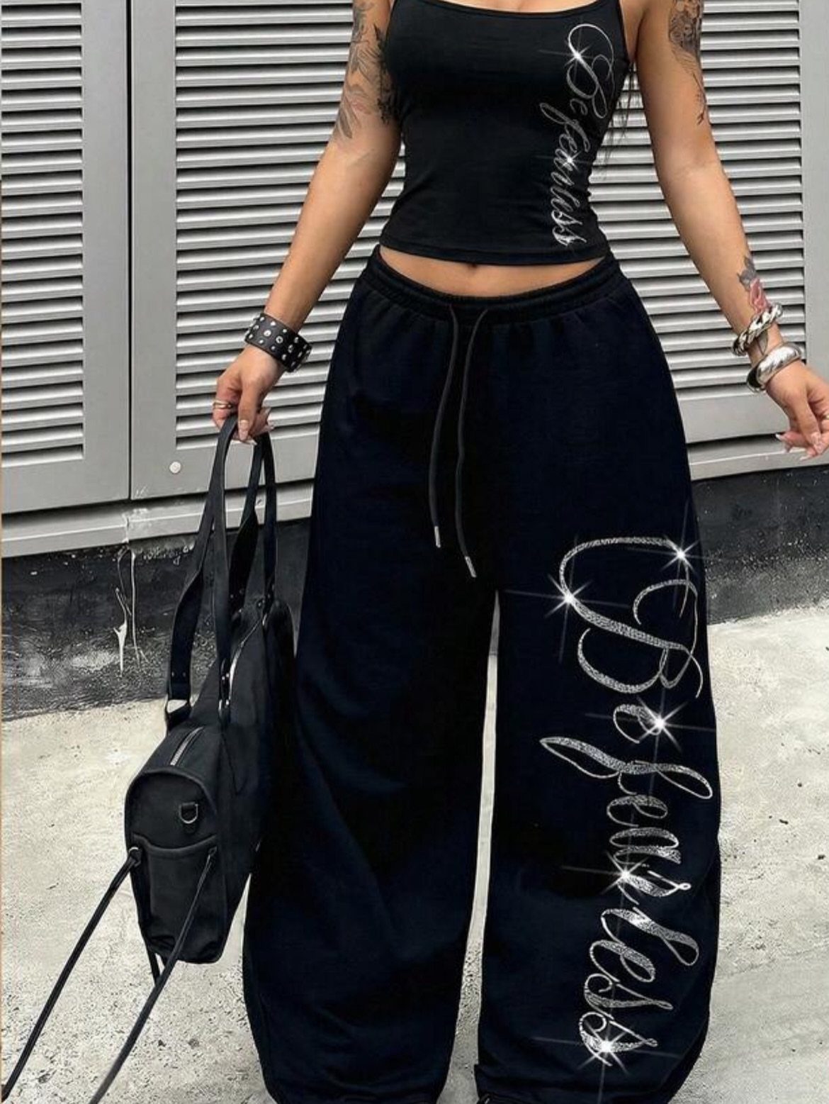 Person wearing a black fitted tank top and baggy joggers with rhinestone lettering.