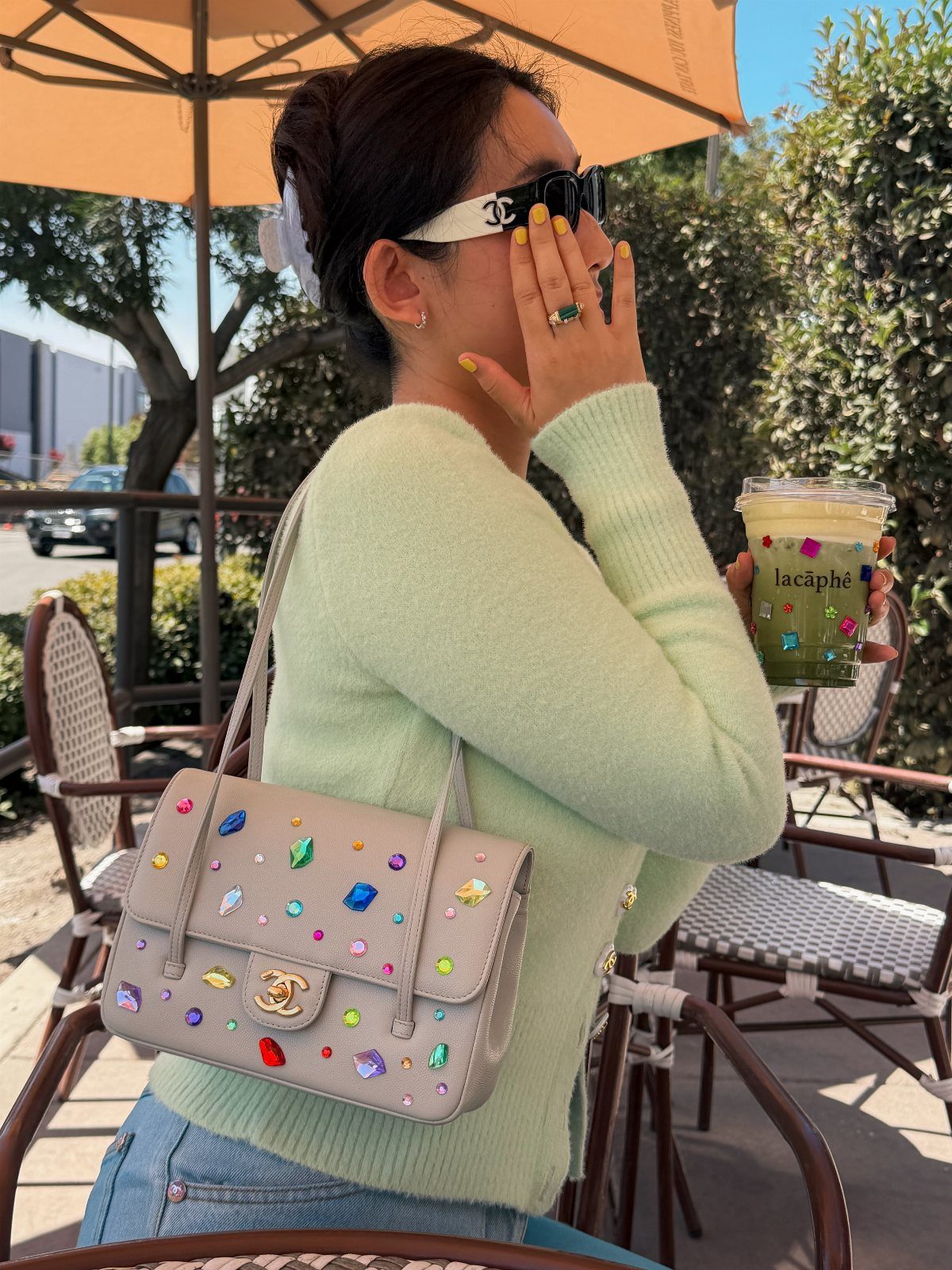 Woman in a green sweater wearing logo sunglasses and holding a rhinestone-embellished handbag outdoors.