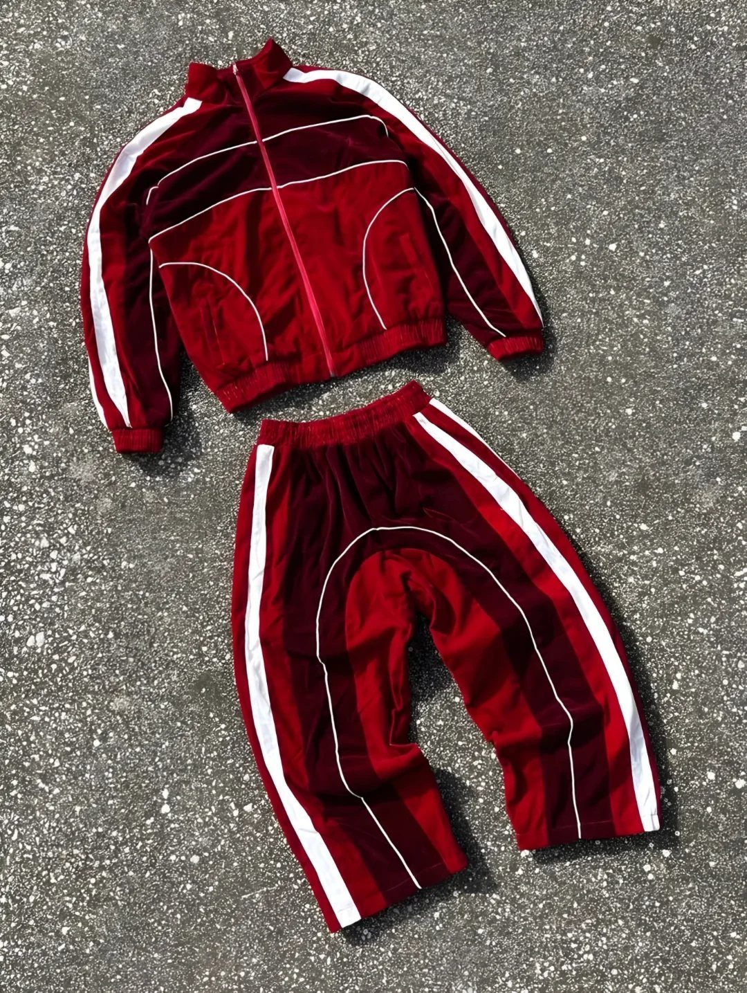 Red velour tracksuit with white stripes laid flat on a grey asphalt ground.