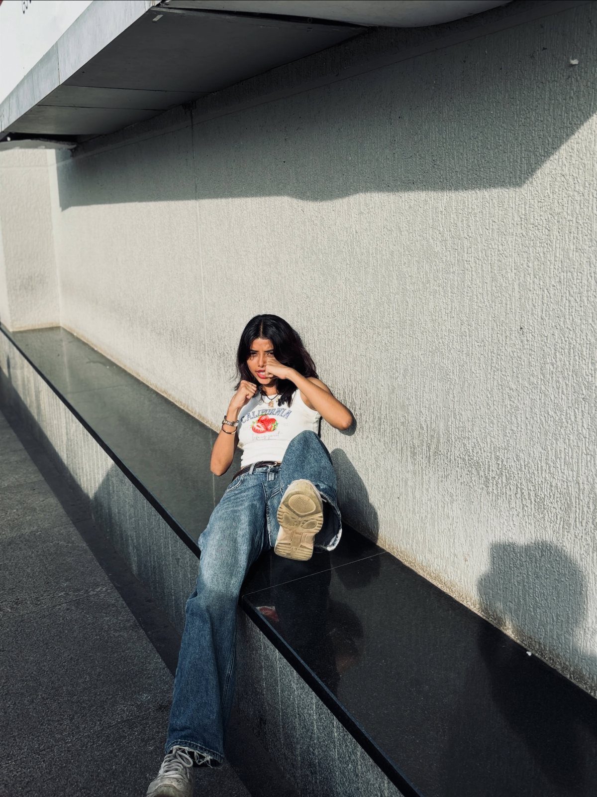 A woman in a white graphic tank top and baggy denim jeans sitting on a ledge.