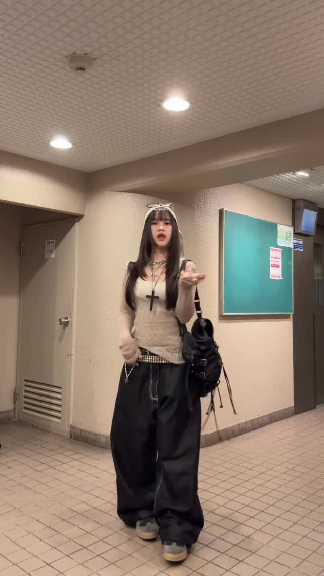 A young woman posing in a hallway wearing baggy black pants and a cream top.