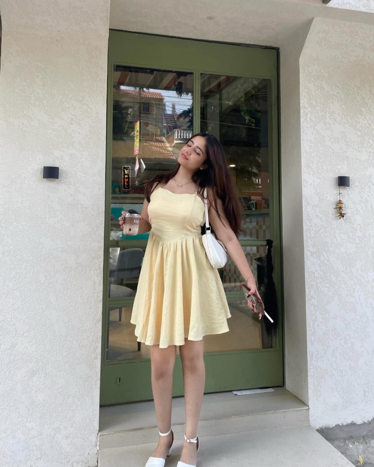 A woman in a pale yellow sleeveless mini dress posing in front of a green door.